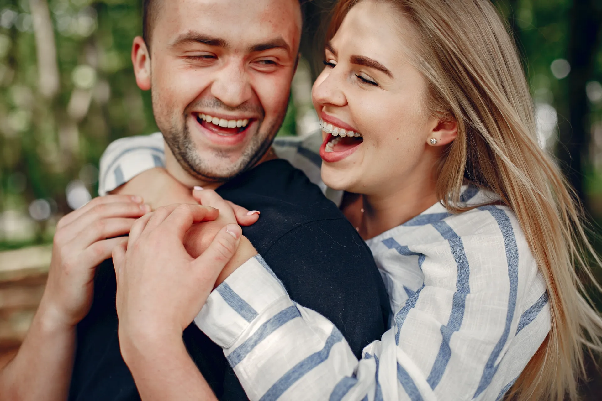 Smiling woman with braces hugging a laughing man from behind outdoors with blurred trees in background.