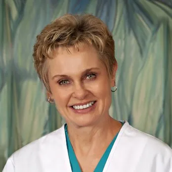 Smiling woman with short blond hair wearing a white medical coat and teal scrubs.