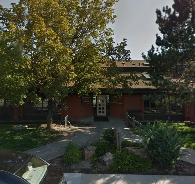 Entrance of a brick building shaded by large trees and surrounded by bushes and a sidewalk.