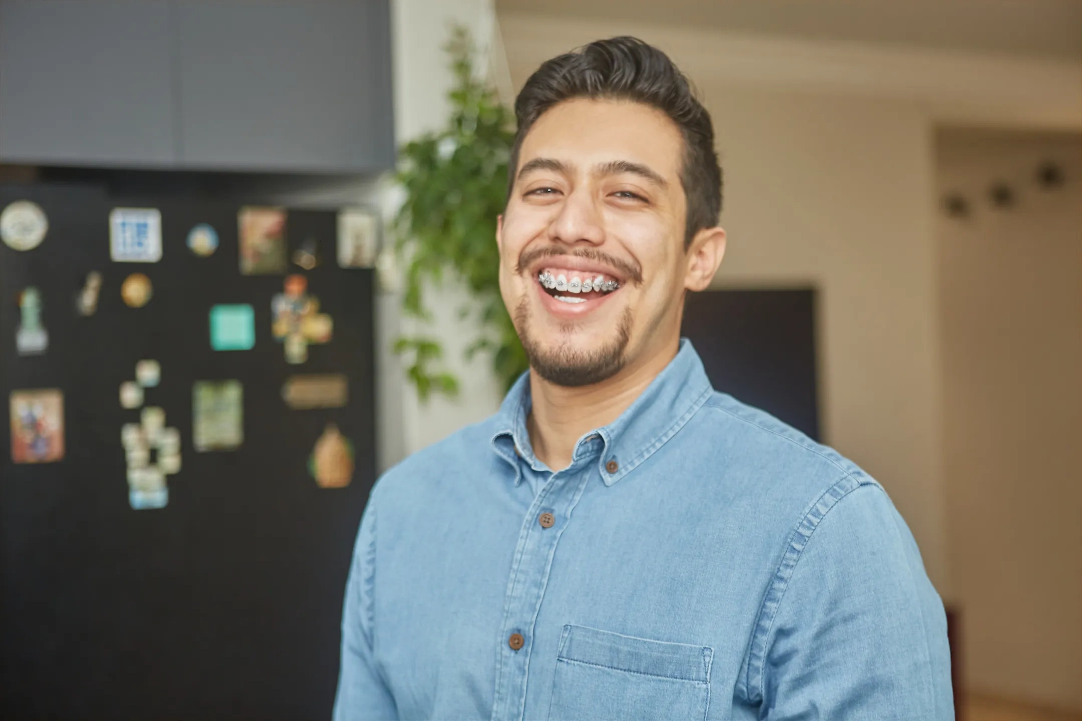 Smiling man with braces wearing a blue buttoned shirt indoors in front of a refrigerator and a plant.