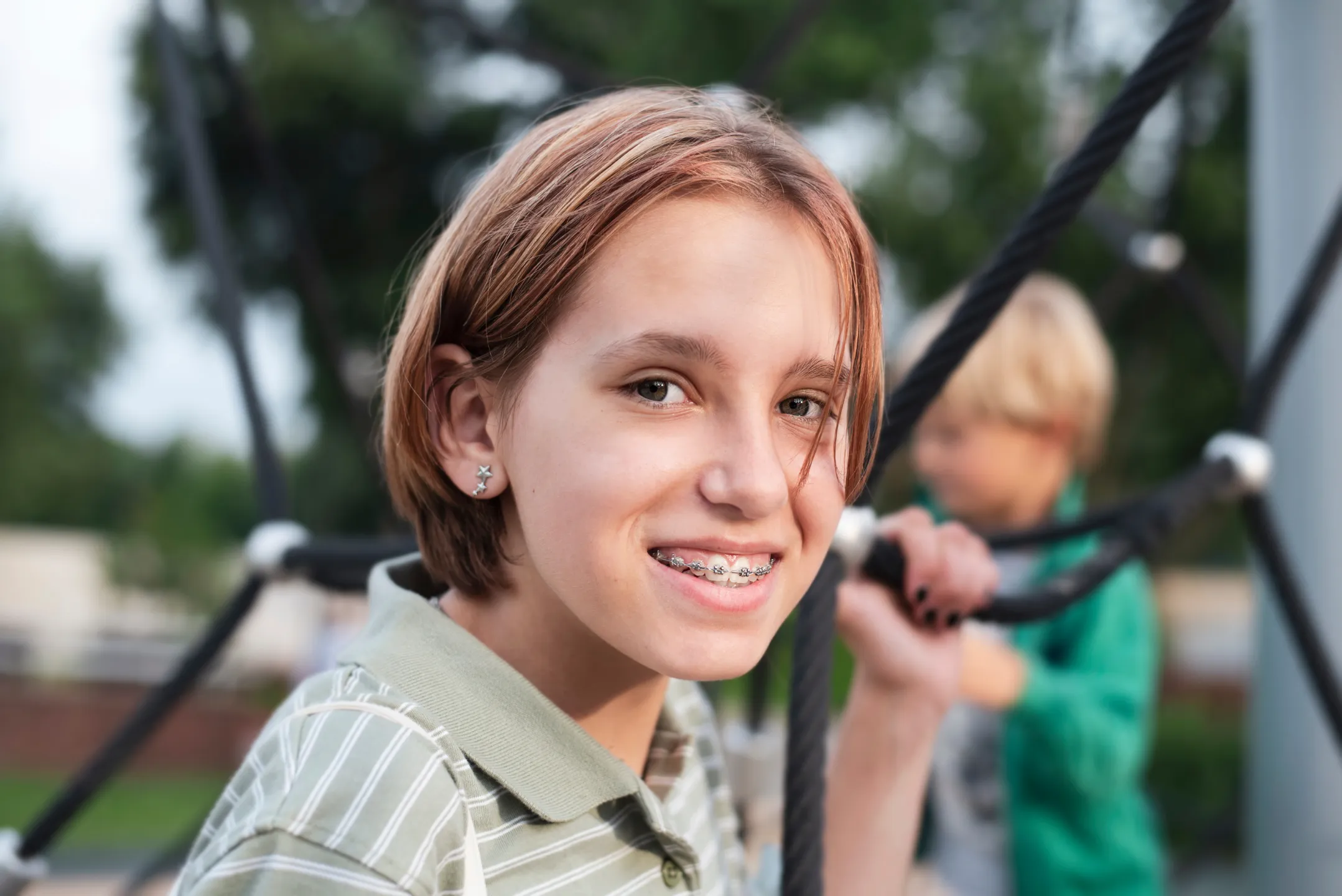 Smiling girl with braces on playground equipment, boy in green jacket in blurred background.