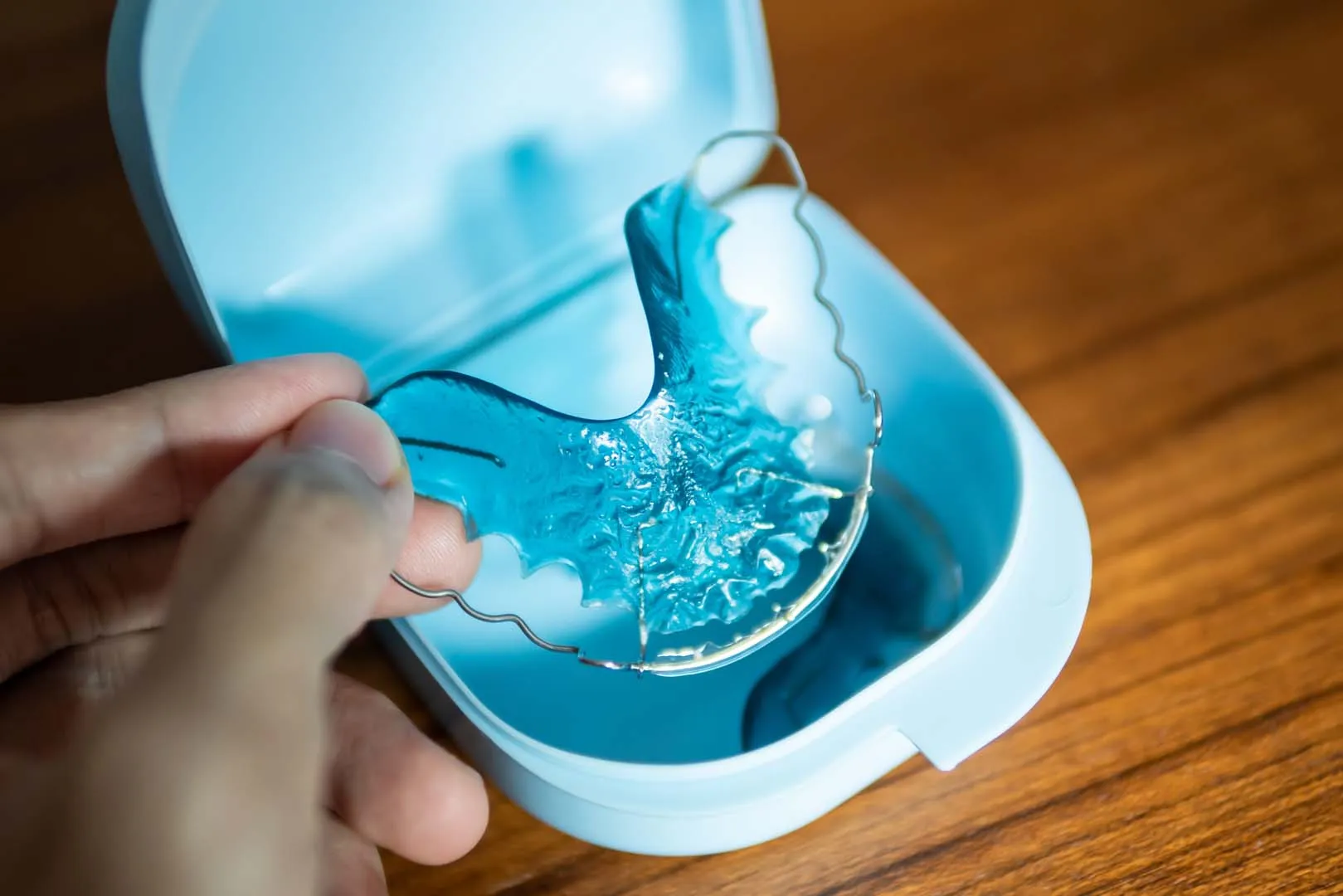 Hand holding blue dental retainer above its light blue plastic case on wooden surface.