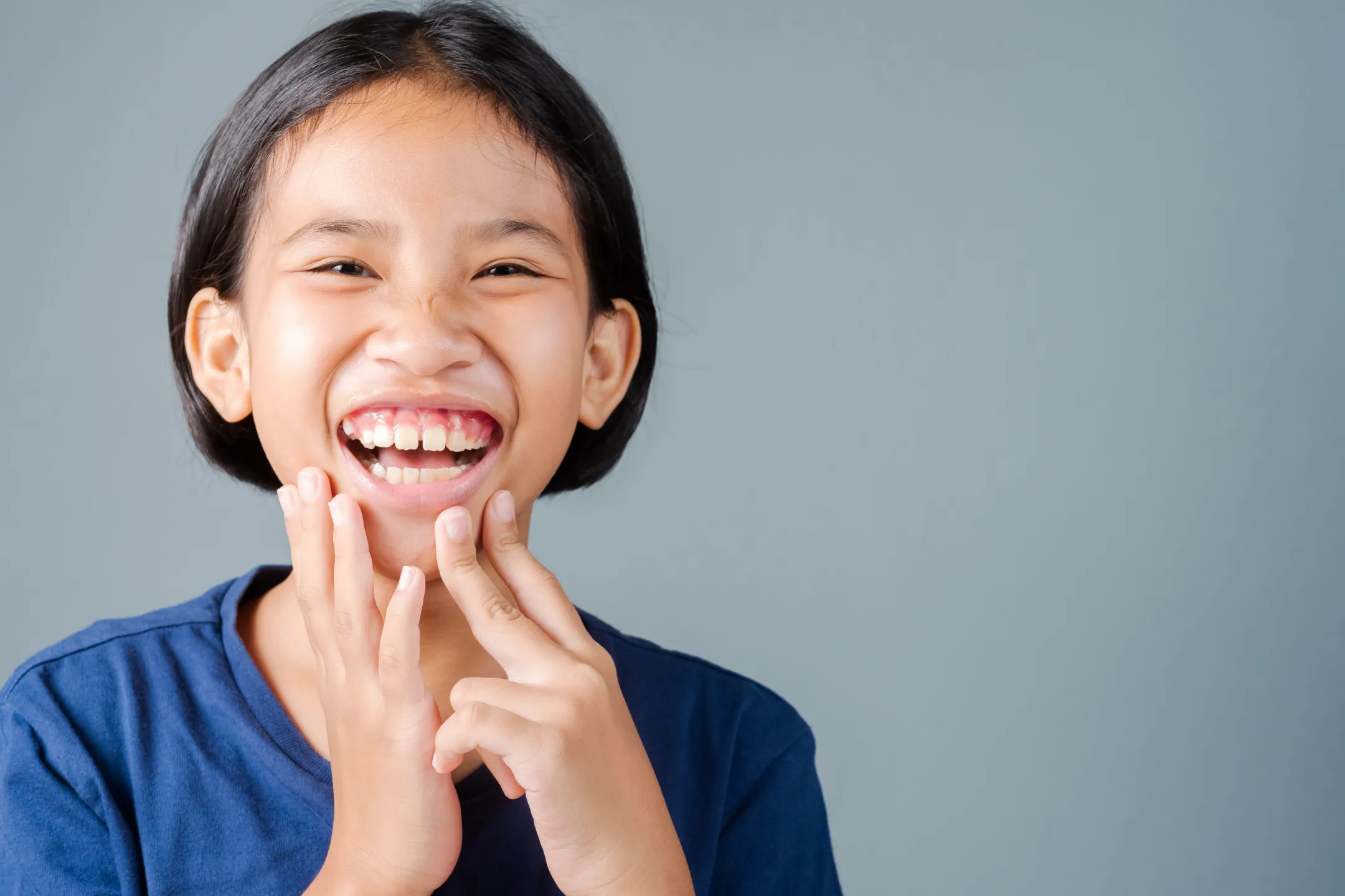 Smiling young girl with short hair shows missing front teeth wearing a blue shirt against gray background.