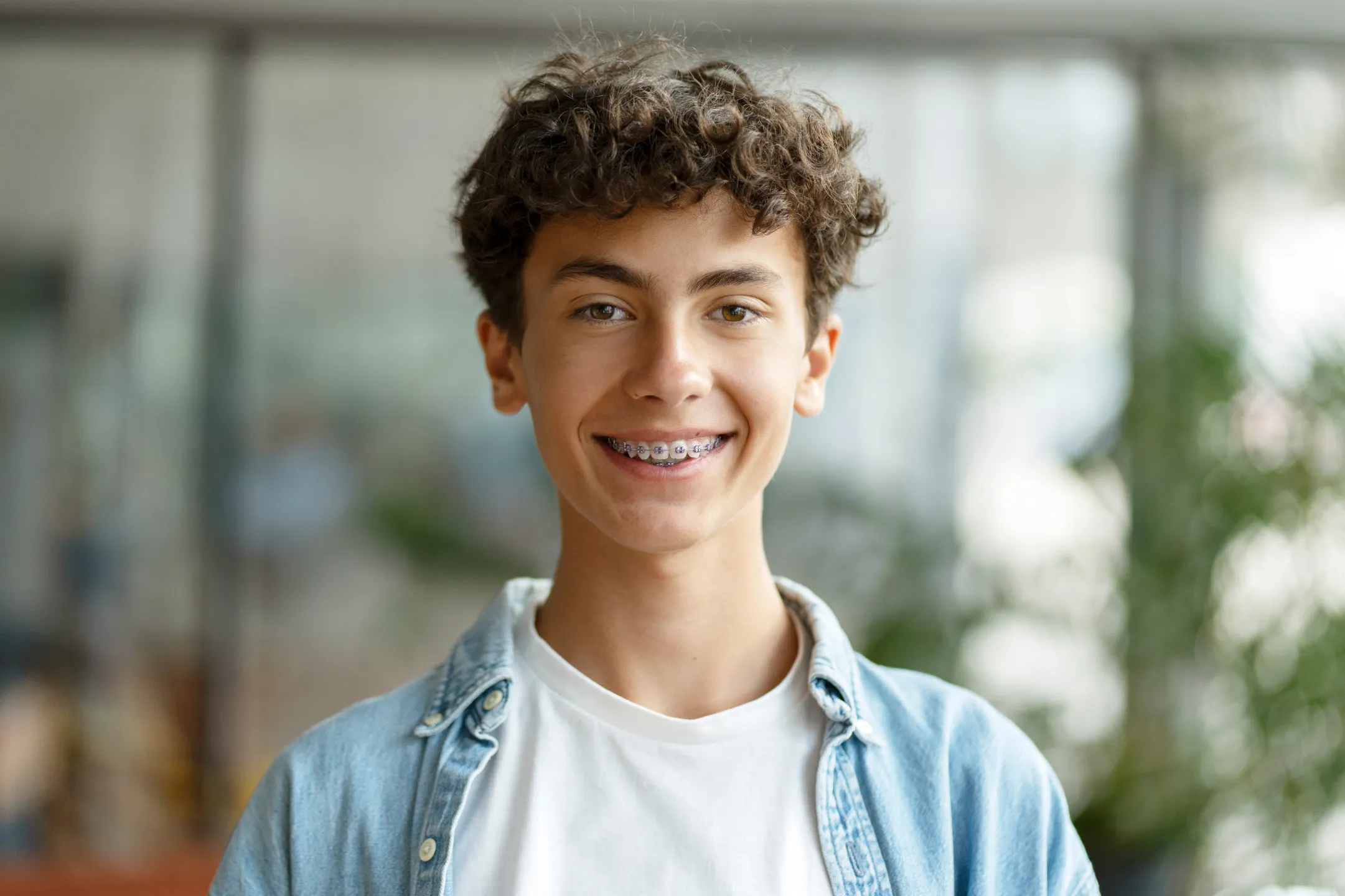 Teen boy with curly hair smiling and showing braces, wearing a white shirt and denim jacket.