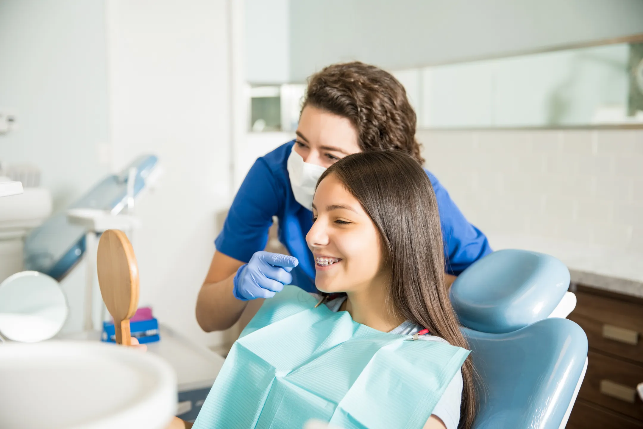 Dentist in mask and gloves showing a teenage girl with braces her teeth in a hand mirror.