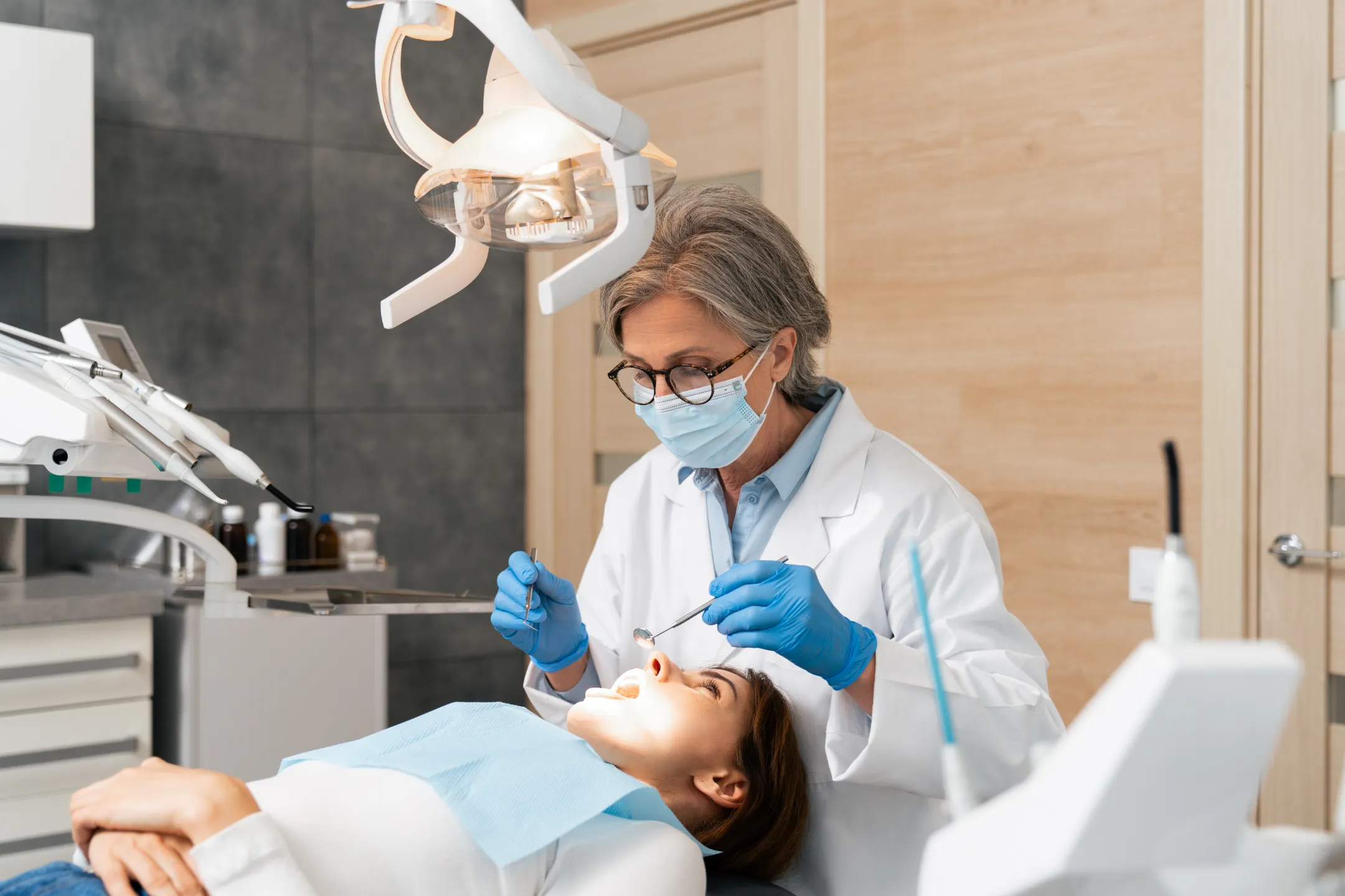 Dentist wearing mask and gloves examining a female patient’s teeth in a dental clinic chair.