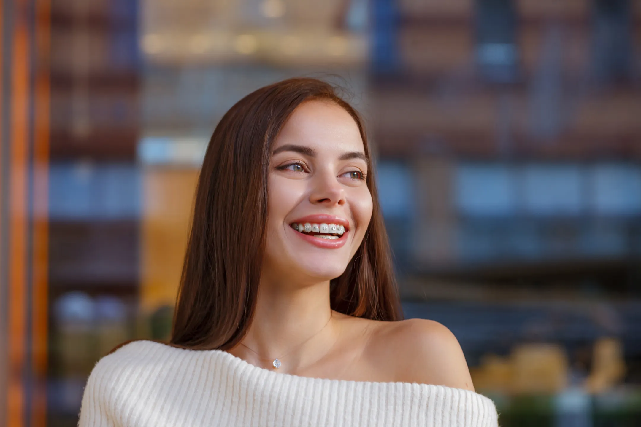 Smiling young woman with braces wearing a white off-shoulder sweater and a necklace.