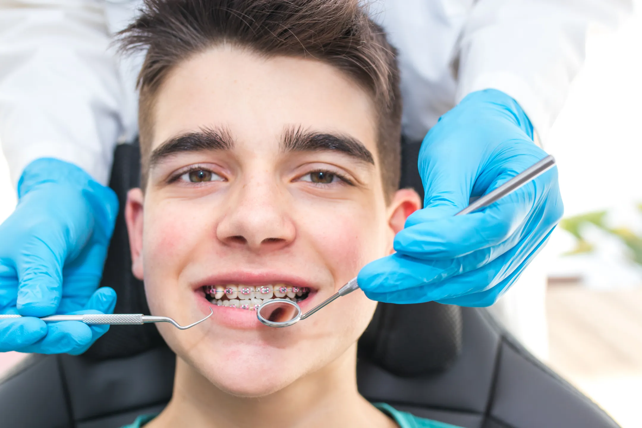 Teen boy smiling with braces as dentist wearing blue gloves holds dental mirror and probe near his mouth.