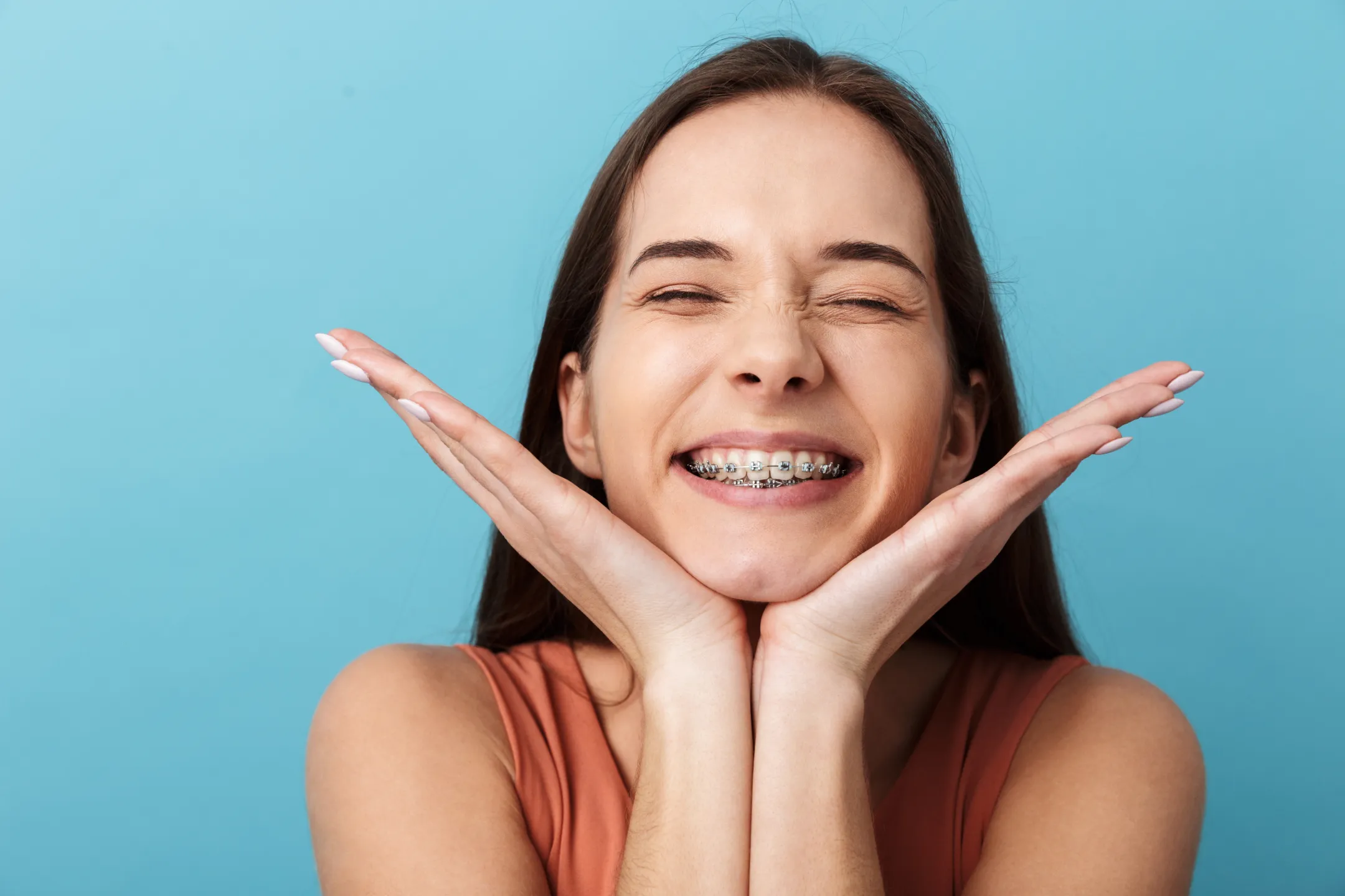 Smiling young woman with braces holding her face with both hands against a blue background.