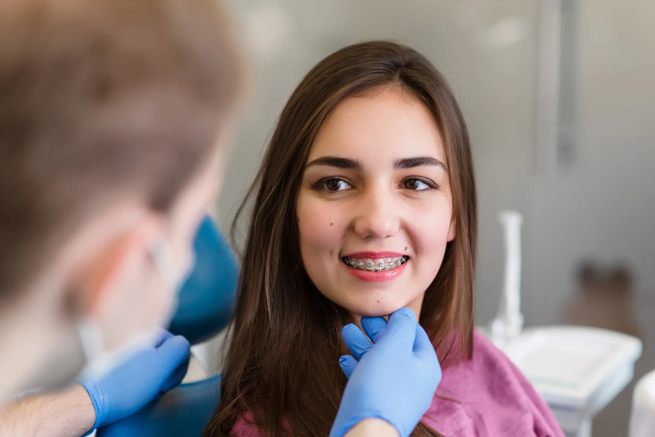 Young woman with braces smiling as dentist wearing blue gloves examines her chin.
