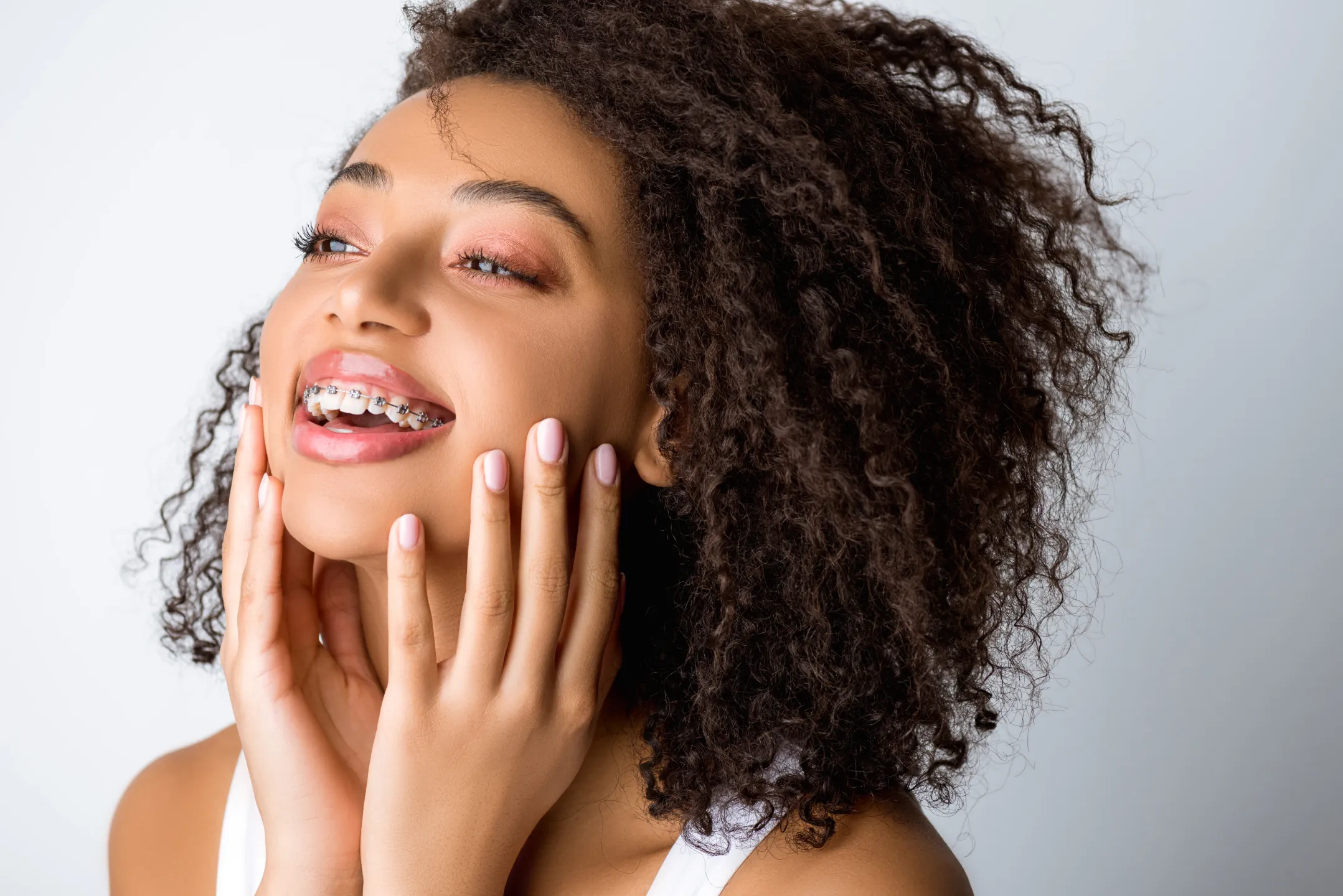 Smiling woman with braces touching her face, showing glossy lips and curly hair.